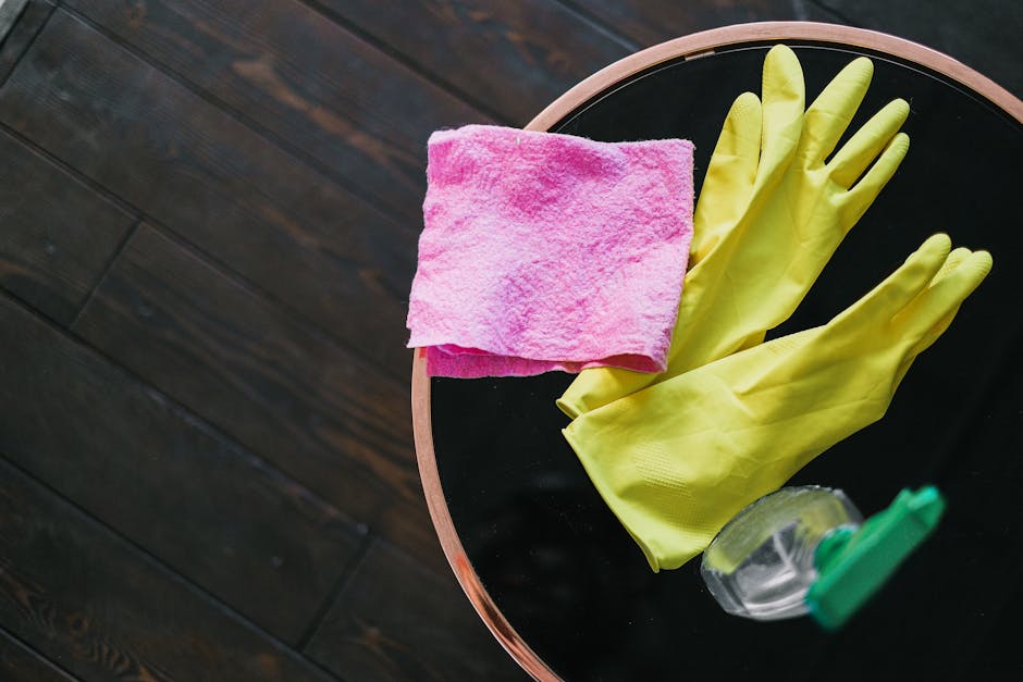 A round black surface with a beige rim, placed on a dark wooden floor, features cleaning tools including a pair of bright yellow rubber gloves, a pink microfiber cloth, and a small spray bottle with a