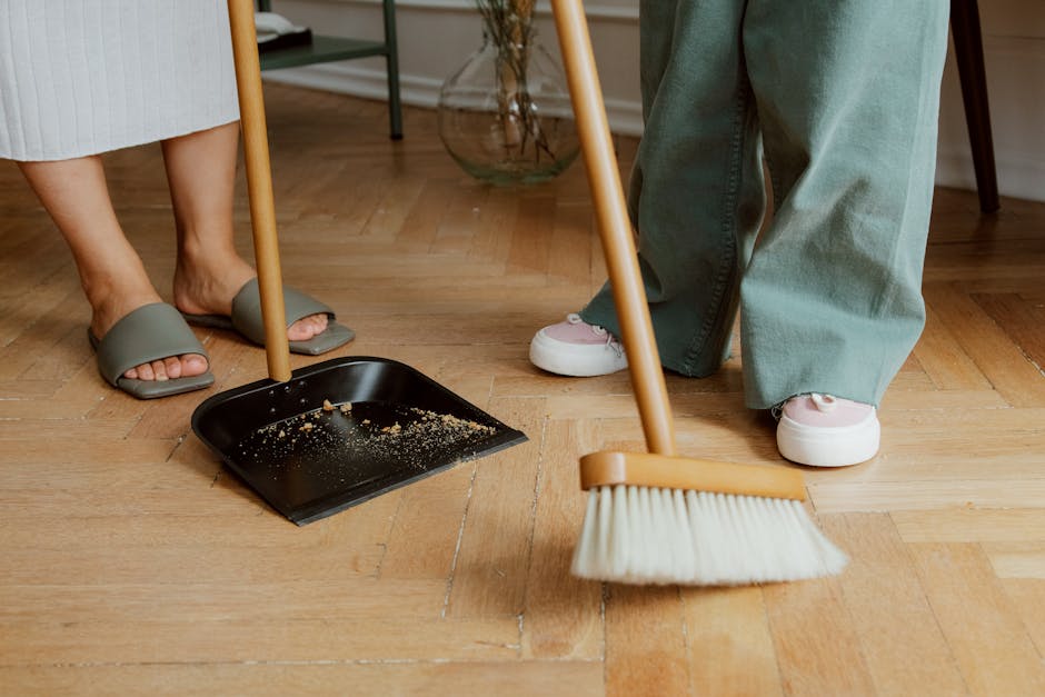 A round black surface with a beige rim, placed on a dark wooden floor, features cleaning tools including a pair of bright yellow rubber gloves, a pink microfiber cloth, and a small spray bottle with a