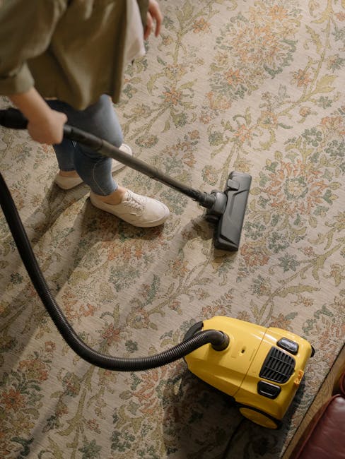 A person wearing beige sneakers and blue jeans is using a vacuum cleaner with a long wand attachment to deep clean a traditional patterned rug in a room. The rug features intricate floral designs in m