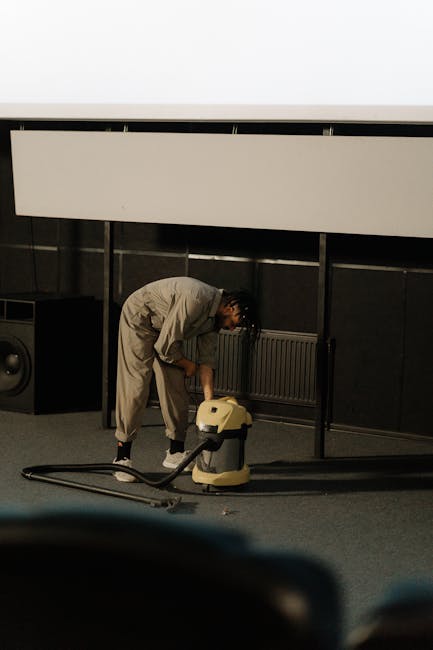 A person wearing beige sneakers and blue jeans is using a vacuum cleaner with a long wand attachment to deep clean a traditional patterned rug in a room. The rug features intricate floral designs in m