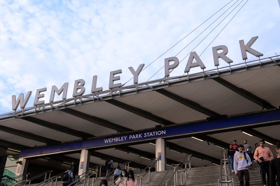 The image shows the interior of Wembley Stadium with a large crowd of spectators seated in the stands, many of whom are holding up their phones or hands. The wide view captures the expansive seating a